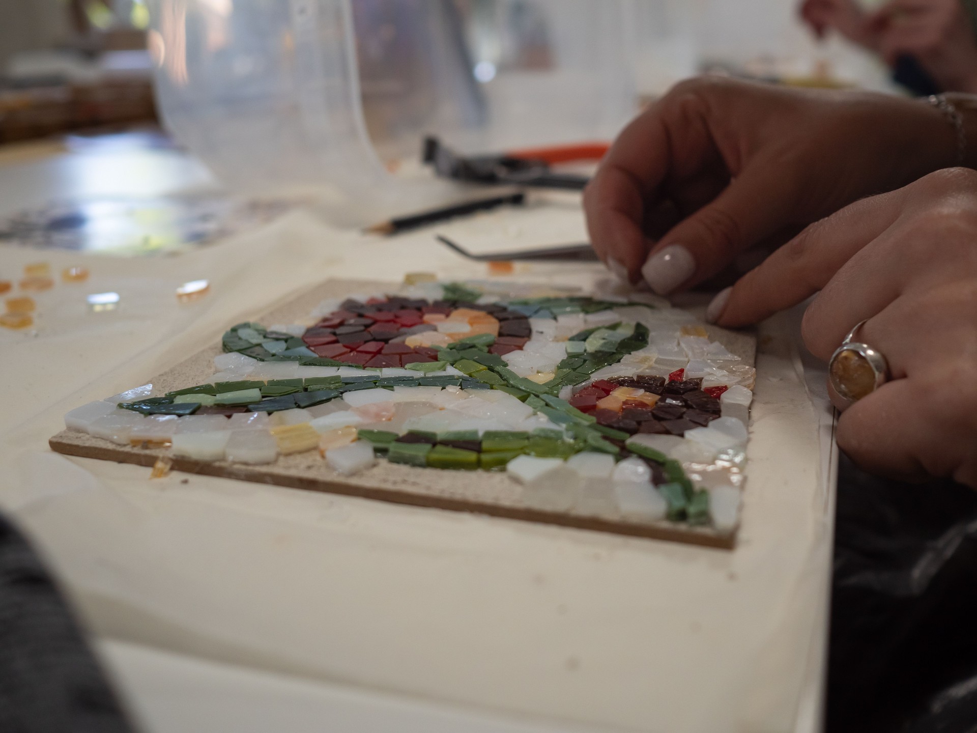 Mosaic Workshop: A horizontal tabletop close-up shot captures a woman's hands crafting a floral mosaic pattern.